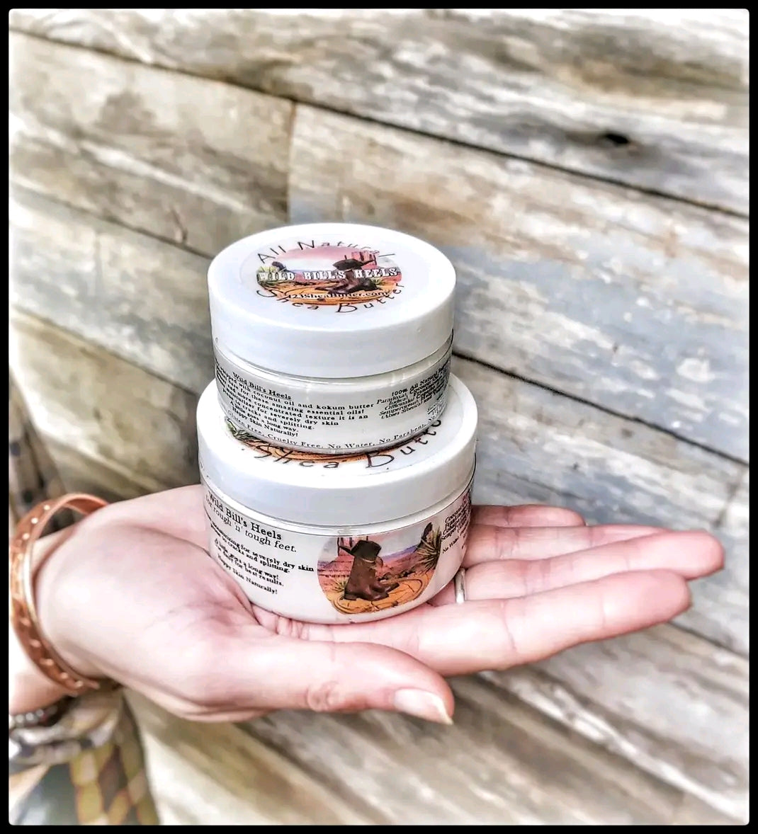 A 4oz and 2 oz jar of Wild Bill's Heels Blend Shea Butter sitting on a hand with a wooden background.