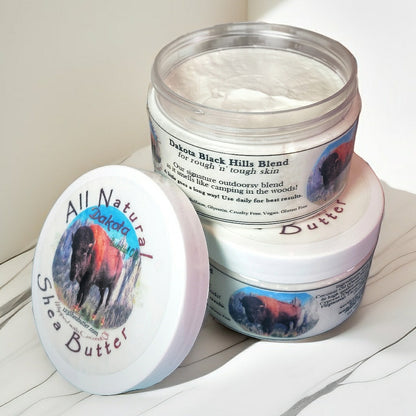 A jar of Dakota Black Hills Shea Butter with the lid off, showing the product inside. The label on the jar features an image of a bison and the text 'All Natural Shea Butter'.