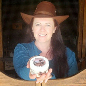 Rebecca wearing a cowboy hat holding a Wild Bill's Heels jar standing behind a wooden railing.