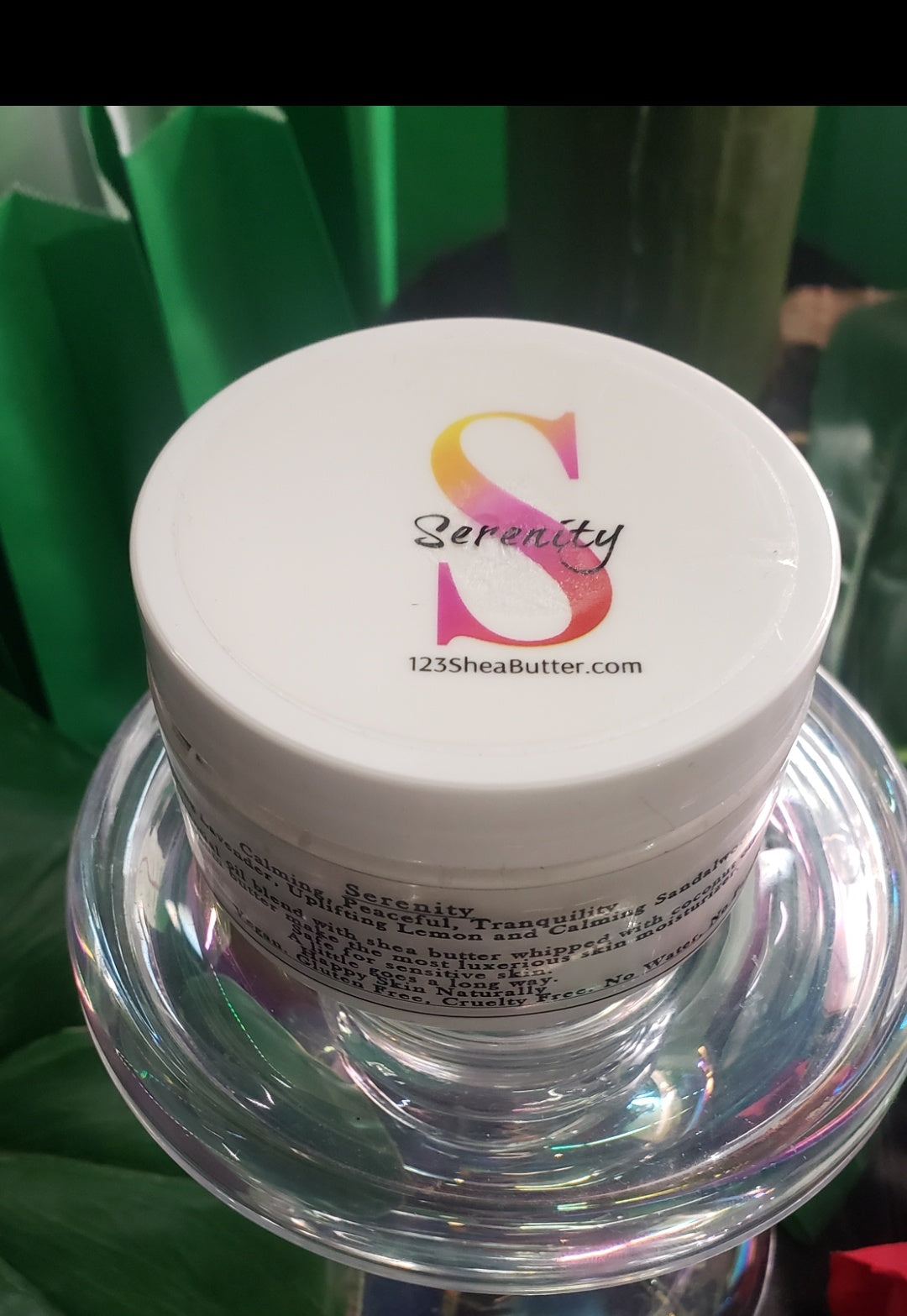 A jar of Serenity shea butter cream with a logo on the lid, placed on a clear pedestal with green background