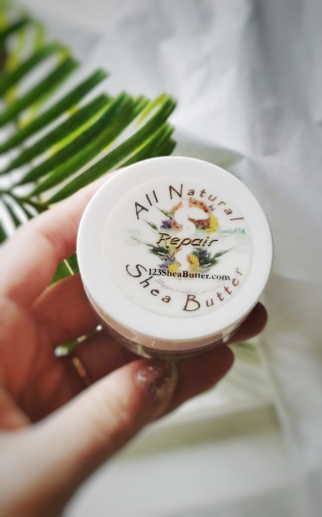 Hand holding a 2oz jar of All Natural Repair Shea Butter with a blurred green leaf background